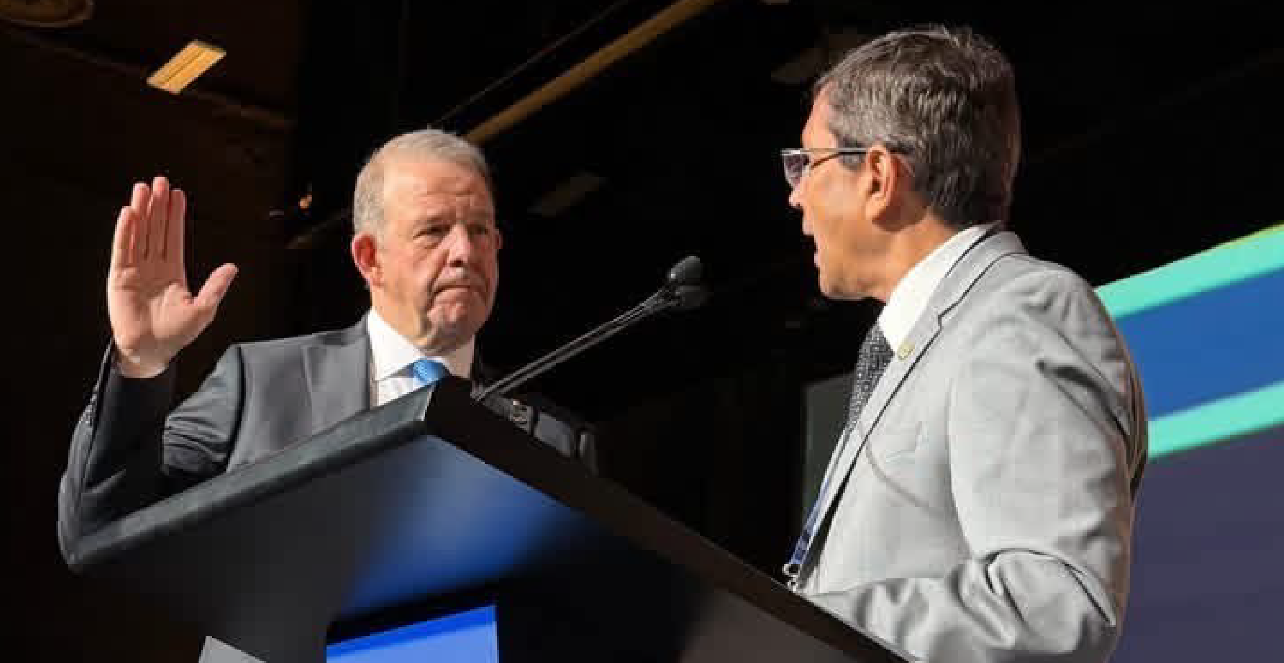 Two men at a podium, one raising his hand in a gesture of oath-taking. Both are dressed in suits, conveying a formal, official atmosphere.