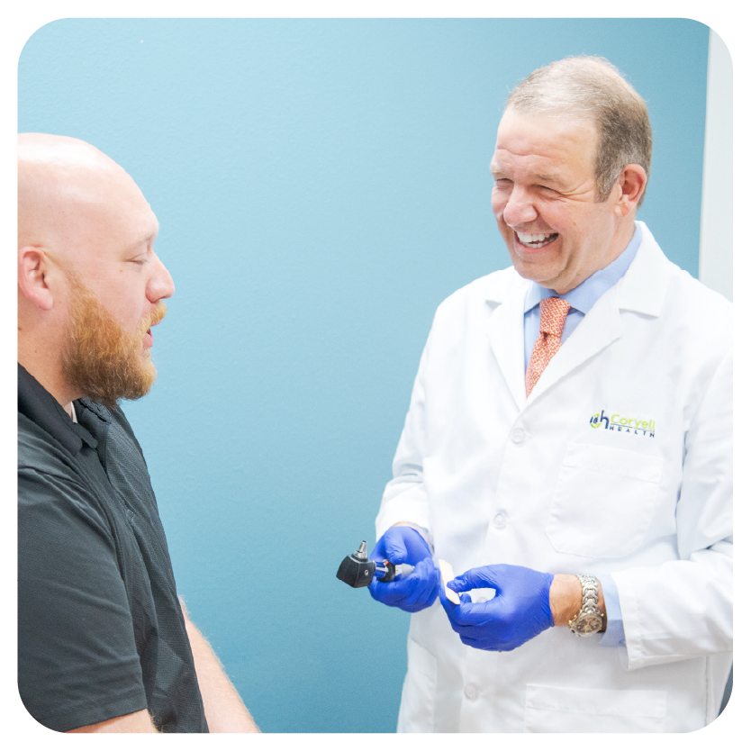 A doctor in a white coat and blue gloves smiles while holding a medical instrument, interacting warmly with a bearded patient, creating a friendly atmosphere.